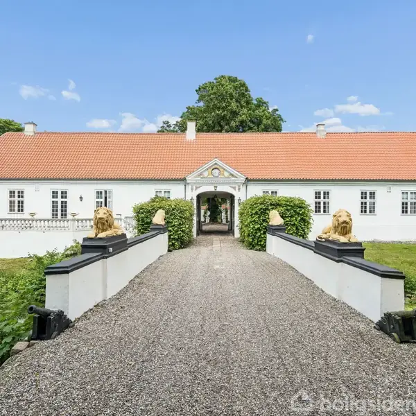 En hvid herregård med rødt tegltag står stille, omkranset af frodige træer. En grusvej fører over en bro med gyldne løvestatuer i forgrunden.