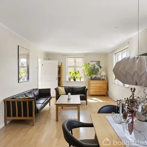 En stue med moderne indretning. Brændeovn til venstre, to sorte sofaer omkring et bord. Et spisebord med stole og en hvid lampe. Lyse trægulve og grønne planter nær vinduer.