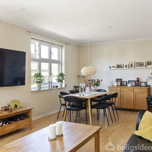 TV hænger på væggen i en lys stue ved siden af et vindue med planter. Bord og stole står i midten. Kommode med billeder og dekorationer i baggrunden. Pejs i hjørnet.