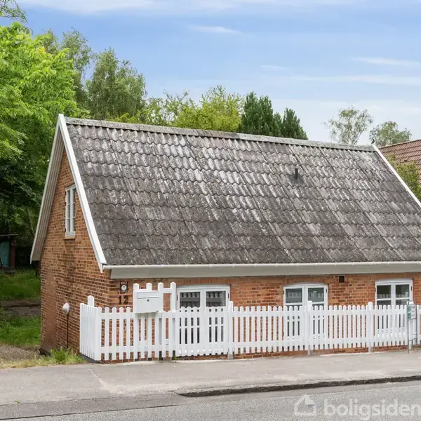 Et lille rødstenshus med et gråt tegltag står bag et hvidt stakit. Huset har tre vinduer foran og er omgivet af grønne træer og buske.