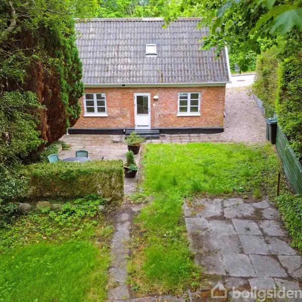Et rødt murstenshus med gråt tegltag står tilbage i billedet. Flisebelagt sti leder gennem en grøn have med høje hække og træer på begge sider.