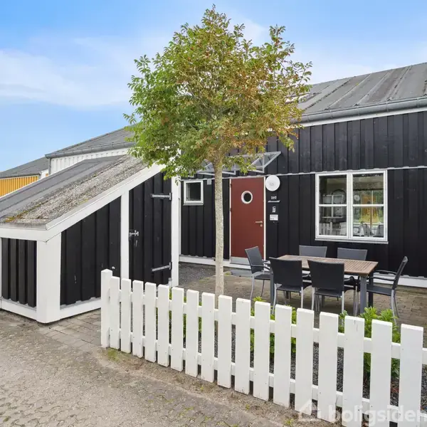 Sort-hvidt hus med hvide hegn, borde og stole foran døren. Det ligger i et velplejet kvarter med fliser og små træer langs vejen.