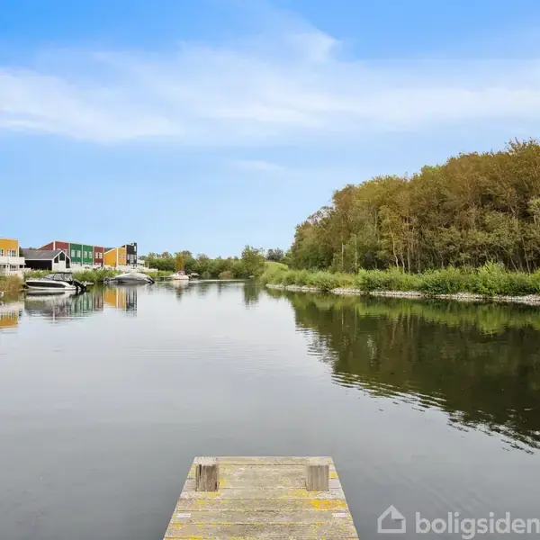 Bro står stille foran en rolig flod, med både fortøjet langs venstre bred. Skov dominerer højre side, og farverige bygninger er synlige i baggrunden under en klar blå himmel.