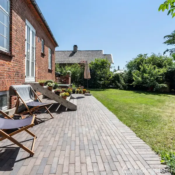 To liggestole står på en flisebelagt terrasse langs en murstensbygning. Haven er frodig med buske og græsplæne, mens en blå himmel danner baggrund.