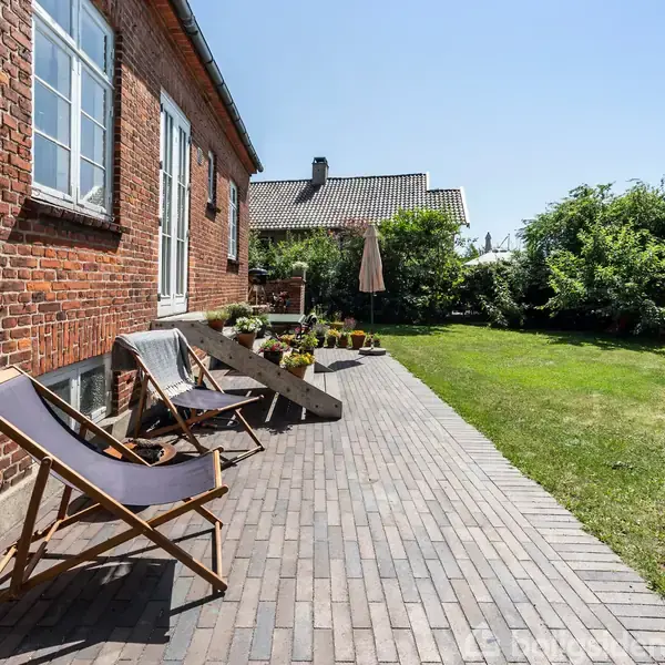 Two lounge chairs stand on a brick patio next to a brick house. Flower pots line the patio steps, and a garden is visible under clear skies.