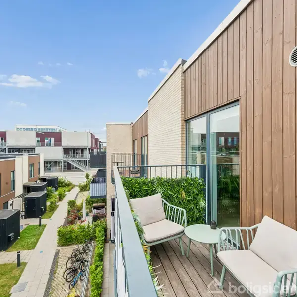 Terrasse med et par stole og et bord står stille. Terrassen er en del af en moderne boligblok, omgivet af grønne områder og cykelstativ. Klar himmel ses i baggrunden.