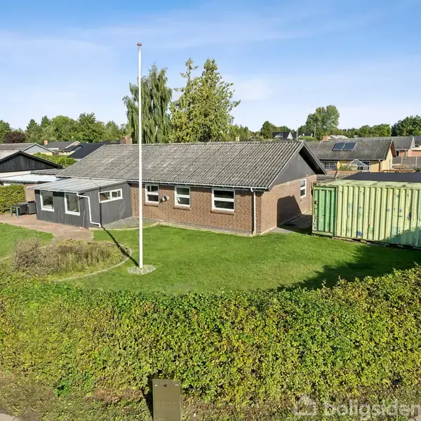 En enkeltstående bygning i mursten står på en velholdt græsplæne. Bygningen har et fladt, gråt tag og er omgivet af en grøn hæk og en flagstang i parcelhuskvarter.