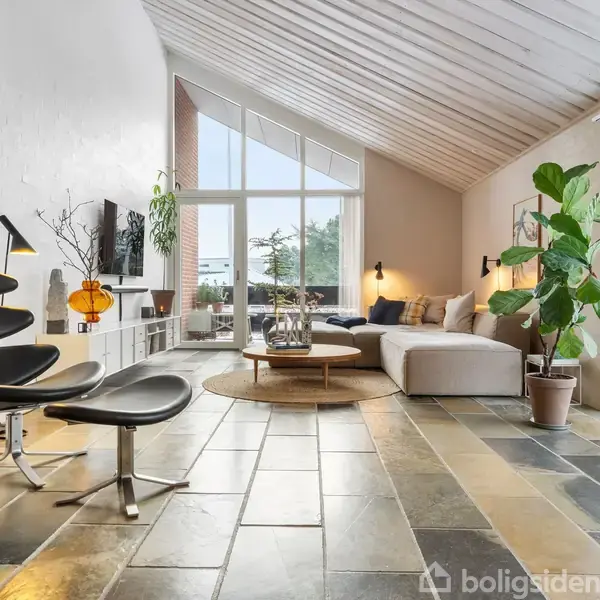 Stue med moderne lænestole og sofabord, stor hjørnesofa og indendørs plante. Handling: Lyst og indbydende rum med store vinduer. Kontekst: Højt til loftet, flisegulv, udgang til terrasse.