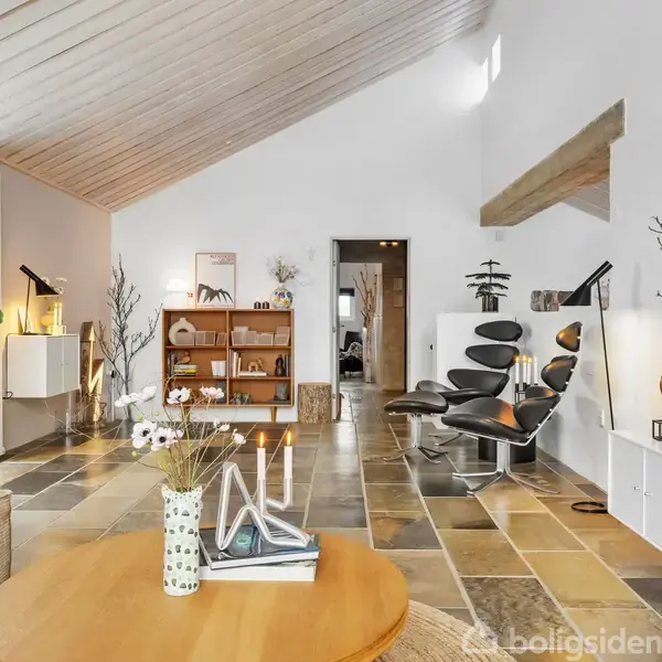 Stue med moderne møblering, herunder en lænestol og et bord med stearinlys. Hylder og planter skaber en hyggelig atmosfære. Rummet har et skråt loft og stengulv.