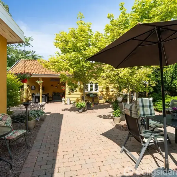 En terrasse med havebord og stole står under en parasol. Omkring er der gule mure, grønne planter og blomster, hvilket skaber en hyggelig haveatmosfære på en solrig dag.