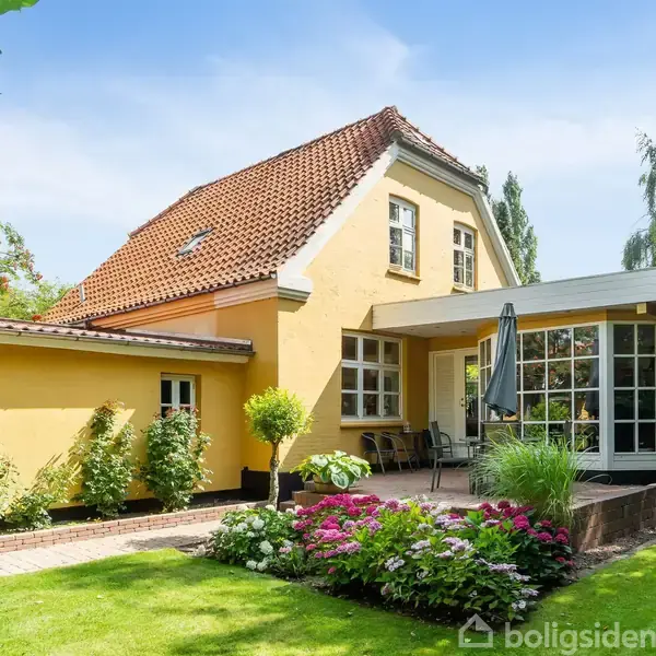 Gult hus med rødt tegltag og en udestue. Terrasse med møbler og parasol. Omkranset af velplejet have med blomsterbede og grønne træer.