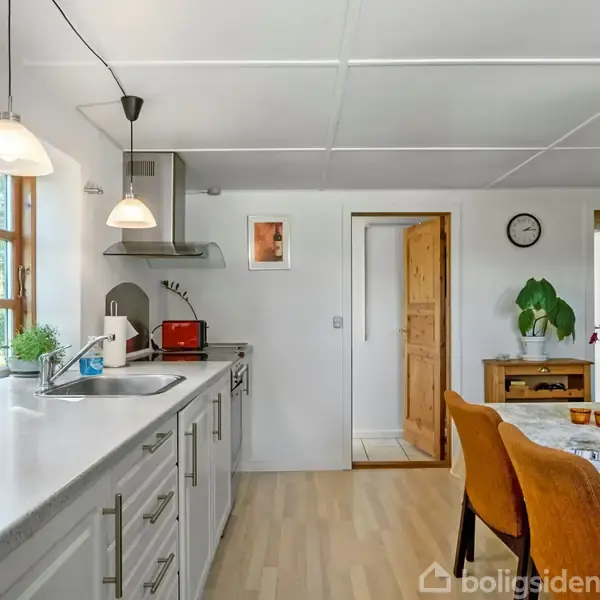 Køkken med hvidtøjvask, stålvask og grå skabslåger, oplyst af hængelamper. Træstole om et bord med blomsterbuket. Væghængt ur og planter i vindueskarmen og hjørne.