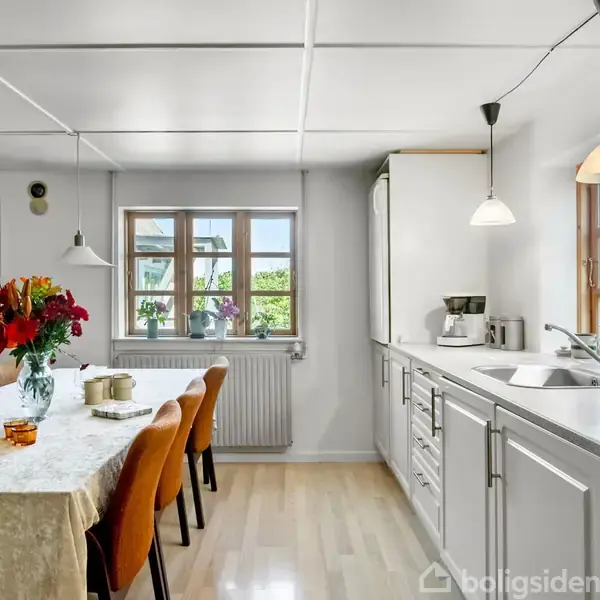 Køkkenbord med blomster og stearinlys står i et lyst rum; hvide skabe og en vaskelinje langs væggen; hængende lamper; vinduer med udsigt til grønne omgivelser.