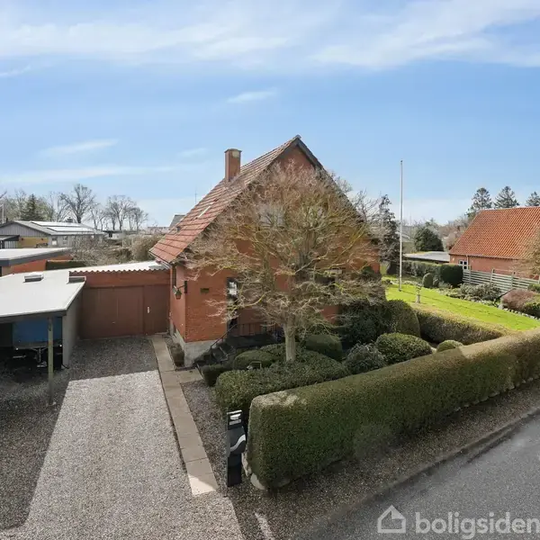 Rødt murstenshus med spidst tag, omgivet af hække og grussti. Carport til venstre. Hyggelig forhave med træer. Beliggende i et boligkvarter med flere huse i baggrunden.