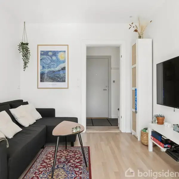 Sort sofa med puder står i en stue ved siden af et lille bord. Rummet har en væg med hængende kunst og et TV. Blomster og planter giver et hyggeligt præg.