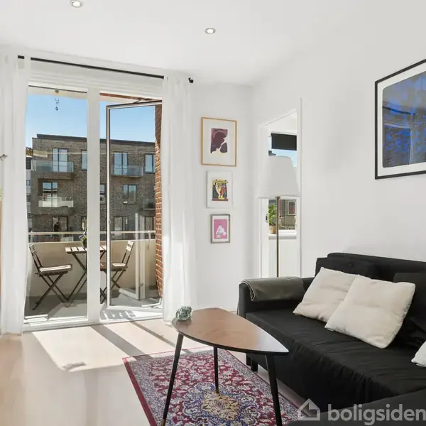 Sort sofa med puder står i en lys stue. En dør åbner til en balkon med et bord og stole. Planter og kunst hænger på væggene.