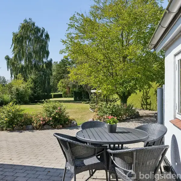 Rundt havebord med fire stole står på en stenbelagt terrasse. Bordet har en lille potteplante. Baggrunden viser en frodig have med græsplæne og høje træer.