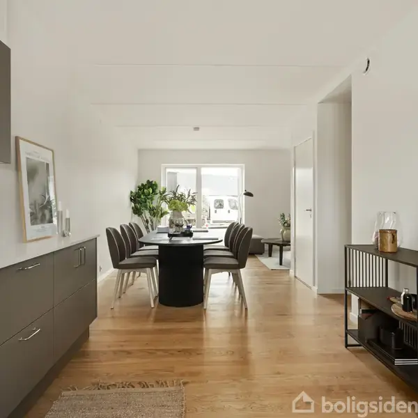 Et moderne spisestuemiljø med et langt bord og stole, omgivet af stilfulde skuffer og skabsenheder. Rummet er lyst med trægulv og grønne planter ved vinduet.