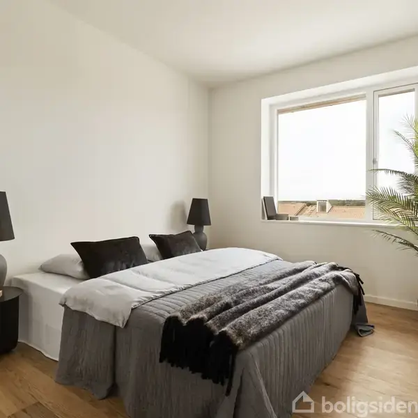 En dobbelt seng med grå sengetøj står i et lyst, minimalistisk soveværelse. To sorte lamper er placeret på natborde, og et stort vindue samt en plante i potte ses i hjørnet.