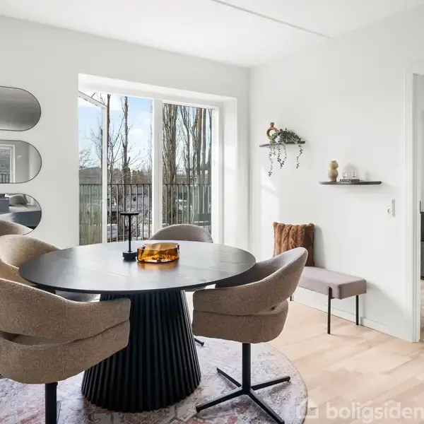 Rundt bord med fire beige stole står i en lys stue, udsigt til balkon med træer. Vægdekorationer omfatter ovale spejle og hylder. Døråbning fører til tilstødende rum.