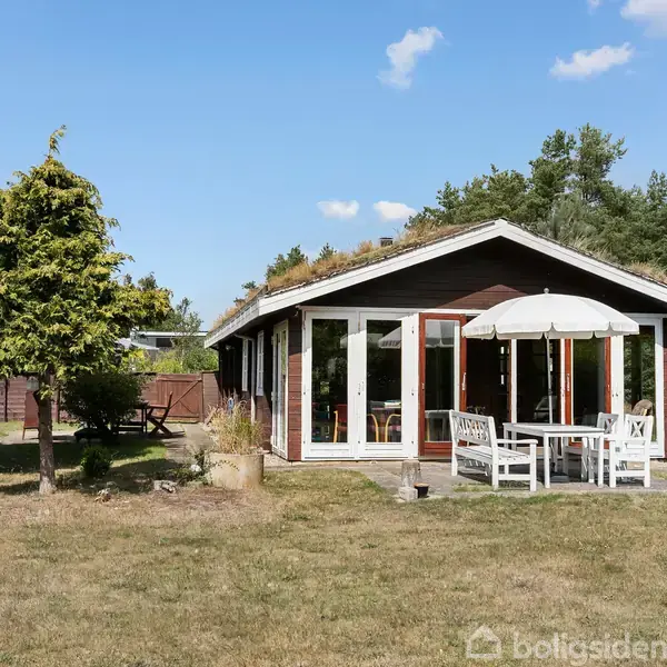 Et lille hus står stille i en grøn have med et bord og stole under en hvid parasol. Træer og buske omkranser bygningen under en blå himmel med få skyer.