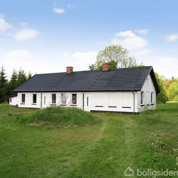 En hvid enfamilieshus med sort tag står stille omgivet af en grøn græsplæne og træer. Huset har flere vinduer og to skorstene, og det er solrigt med blå himmel.