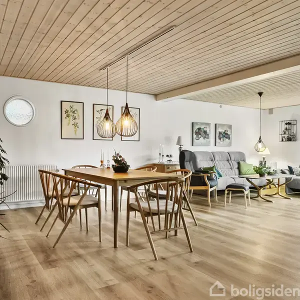 Et spisebord med stole står i et lyst stueområde. Loftet er af træ, og der hænger lamper over bordet. I baggrunden ses en grå sofa og sofabord.