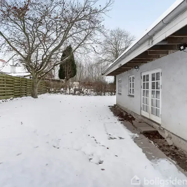 Et snedækket haveområde med et hus til højre. Huset har hvide vinduer og en terrasse i sneen. Der er træer og en træhegn i baggrunden.