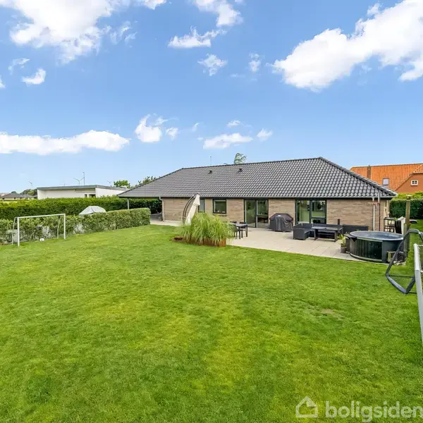 En etplans villa med grå tag sten ligger i en velplejet have med grønne græsarealer, omkranset af hække. Der ses en stor terrasse med havemøbler.