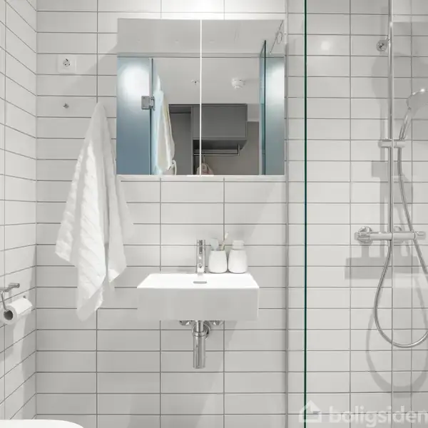 Et moderne badeværelse med hvid flisevæg. En håndvask med spejl og toalettsager ses foran. En bruser er placeret til højre, og et håndklæde hænger til venstre.