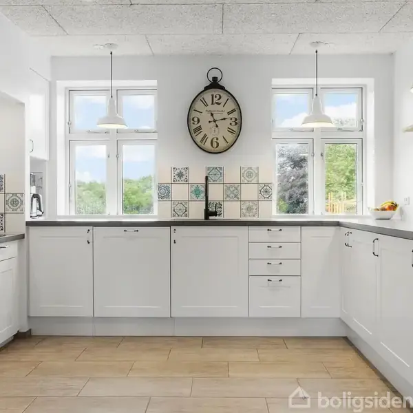 Et lyst køkken med hvide skabe omkranser et åbent rum. Et stort ur hænger over fliseksprydede vægge, mens vinduer lukker naturligt lys ind.