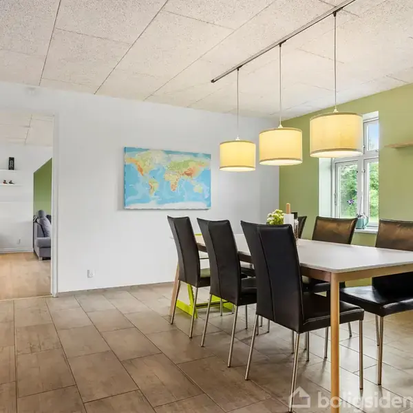 Spisebord med sorte stole står i et lyst rum. Over bordet hænger tre lamper. Kort hænger på væggen, og en hylde med pyntegenstande er ved vinduet.