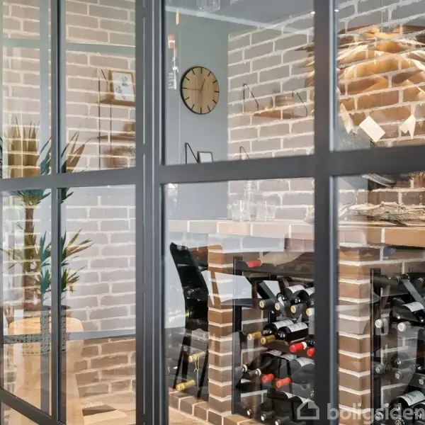 En vinreol fyldt med flasker ses gennem en glasvæg. Rummet har en væg af mursten, en ur på væggen, og en plante til venstre.