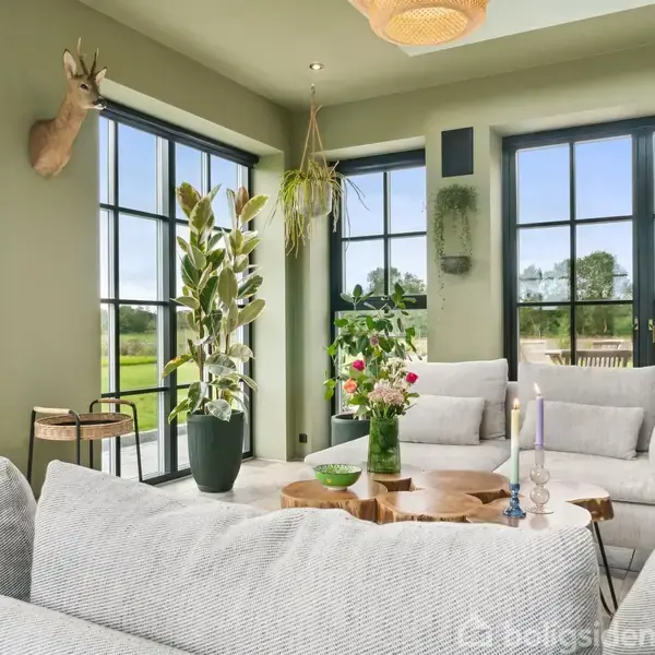 En stue med lysegrå sofaer står foran store vinduer, der lukker naturligt lys ind. Der er en træbord beklædt med en vase fyldt med blomster og levende lys. En hjortebuste hænger på væggen.