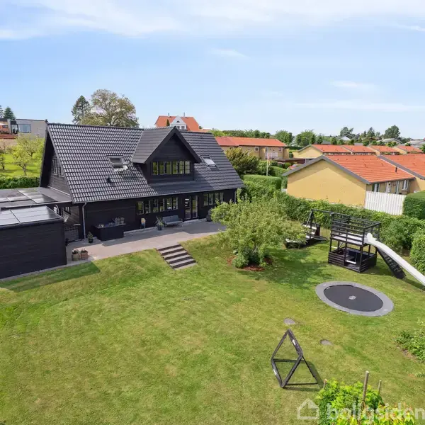 Sort hus med terrasse set bagfra, i en velplejet have med legeområde, rutsjebane, trampolin og gyngestativ. Omgivet af hække og nabohuse i et boligområde.