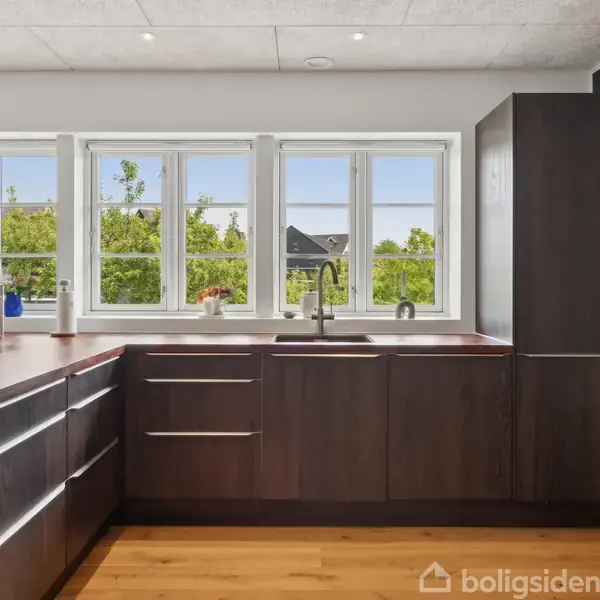 Moderne køkken med mørke træskabe står i en stilfuld indretning. Store vinduer slipper naturligt lys ind, mens en skål med frugt pynter på køkkenbordet. Udsigt udenfor viser grønne træer.