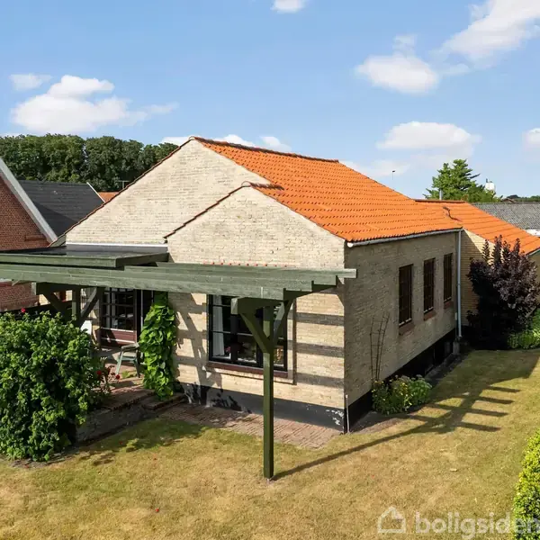 Huset har orange tegltag og lyse mursten, omgivet af en velplejet have med klippede hække og græs. En pergola står ved siden af huset, som ligger i et roligt boligområde.