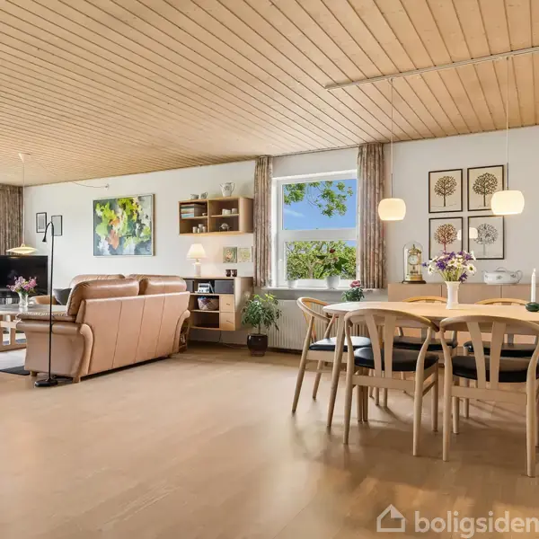 Stue med lyse trægulve og loft, brun lædersofa og spisebord med stole. Kunstværker pryder væggene, mens vinduer viser udsigt til grønne omgivelser. Lamper hænger over bordene.