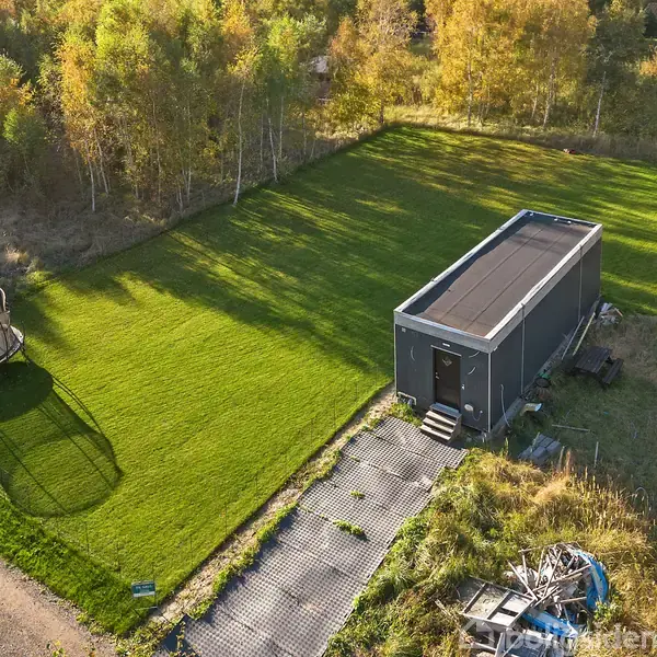 Et lille rektangulært hus med solpaneler står på en velplejet græsplæne. Ved siden af er en trampolin. Omgivet af træer og en sti langs grunden.