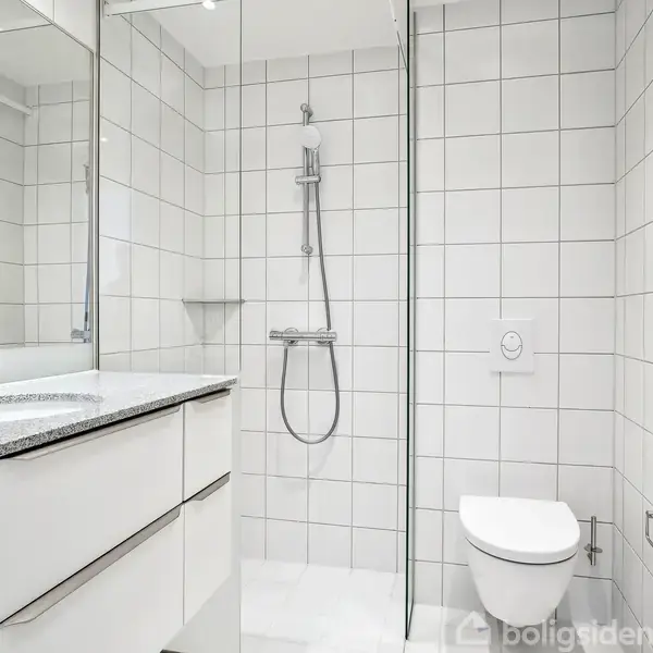 Et moderne badeværelse med et spejl over en håndvask med granitbordplade, brusekabine med glasvæg og væghængt toilet. Rummet er omgivet af hvide fliser og har et rent design.