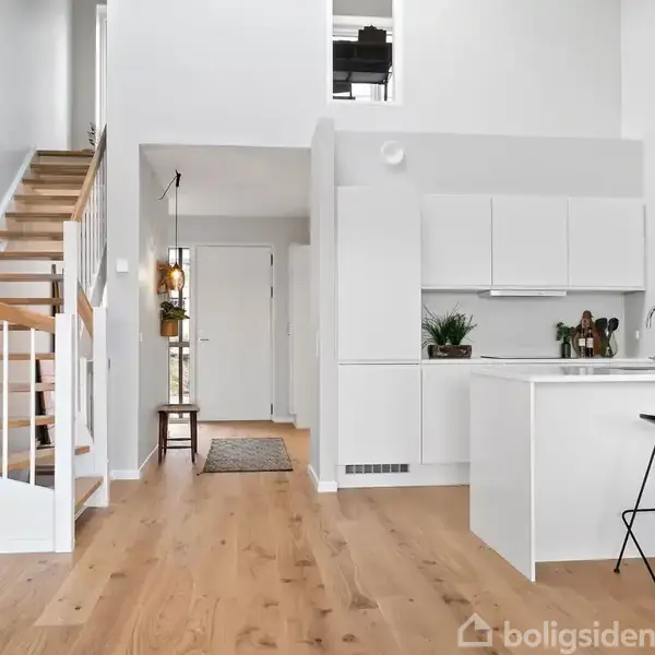 Trappe i lyse træmaterialer stiger op til førstesal, mens moderne køkken med hvide skabe og køkkenø med barstole står til højre. Plantegrønt pynter rummet. Gulvet er i lyst træ.