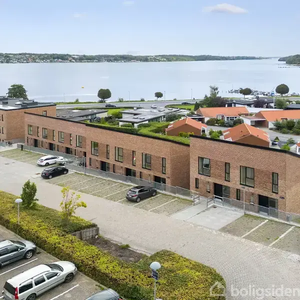Rækkehuse af mursten står langs en vej med parkeringspladser foran. Omkranset af grønt områder, ligger de tæt på en sø med udsigt til træer og bebyggelse på den anden side.