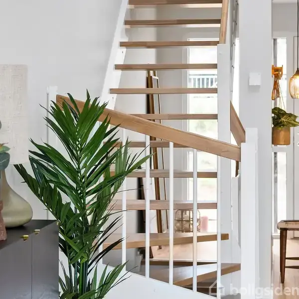 Trappe med trætrin fører opad, omgivet af hvide vægge. Til venstre står en plante og en skulptur på et skab. En lampe hænger over en skammel.