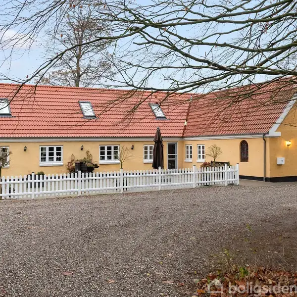 Gult hus med rødt tegltag, flere vinduer og en hvid stakitfront. I gårdspladsen står et sort havebord med parasol. Bladmønstrede trægrene hænger ned fra toppen.