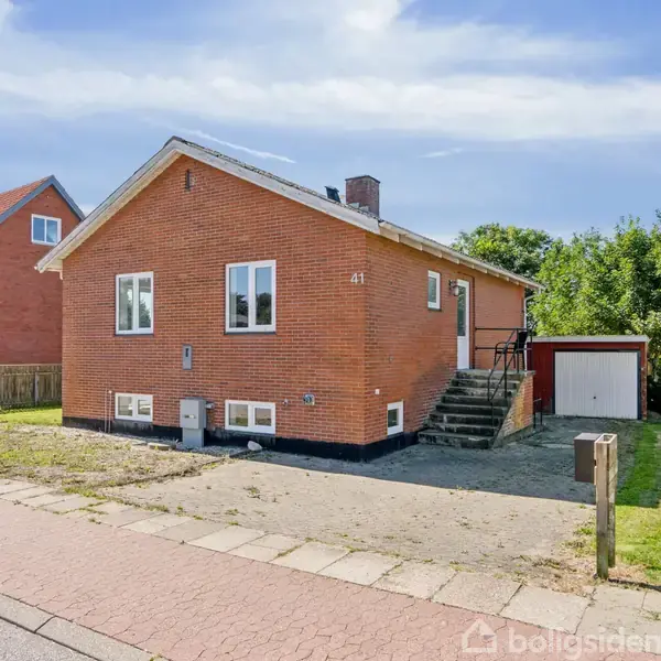 En murstensvilla med nummeret 41 på facaden. Trappen fører op til hoveddøren. Bygningen ligger langs en vejkant, omgivet af græs og få træer i baggrunden.