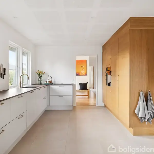 Køkken med hvide skabe, moderne vask og ovn indbygget i en træpanelvæg. Sollys strømmer ind gennem to store vinduer, og der hænger viskestykker på væggen.