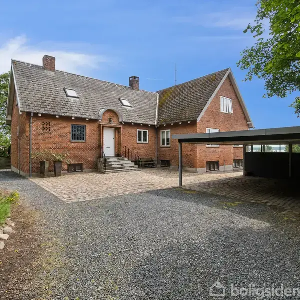 Rød murstensvilla står stille ved en grusbelagt indkørsel, omgivet af træer og grønne planter. En carport i højre side giver skygge, og himlen er klar og blå.