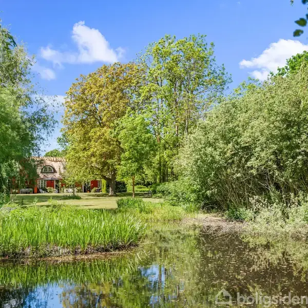 En sø afgrænset af træer og buske reflekterer en klar, blå himmel. I baggrunden ses et stort, rødt hus omgivet af en grøn græsplæne og frodig vegetation.
