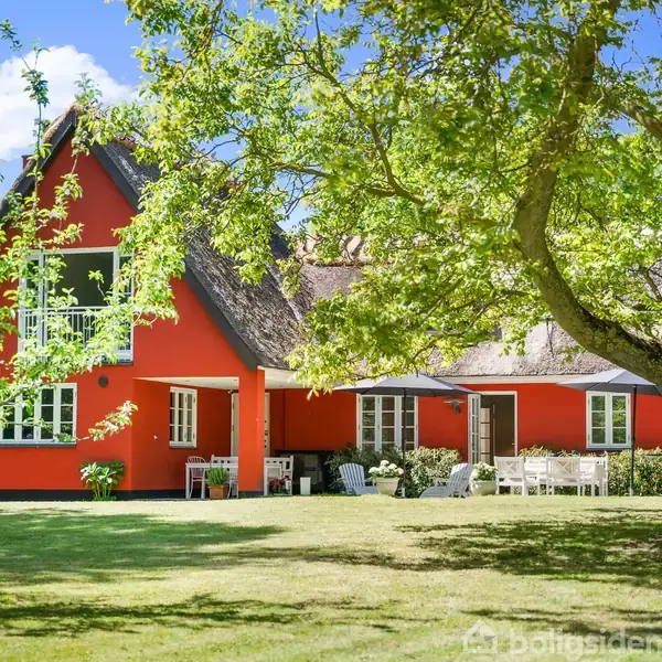 Rødt hus med stråtækt tag står stille i en grøn have omkranset af træer under en blå himmel. Der er ingen synlig tekst i billedet.