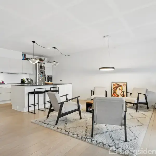 Stue med moderne møbler i lyse farver, hvor to stole og et sofabord står på et tæppe. Køkken til venstre er åbent og minimalistisk. Vægbillede tilføjer kunstnerisk touch.